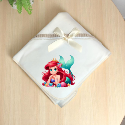 Toalha de bebé branca com laço creme e estampa de sereia colorida sobre mesa de madeira clara com planta ao lado