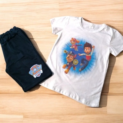 Conjunto infantil de t-shirt branca com personagens da Patrulha Pata e calças pretas, dispostos sobre chão de madeira clara.