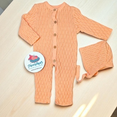 Macacão de bebé cor de pêssego com padrões trançados e botões de madeira, com touca igual e etiqueta PIRRALHOS sobre mesa clara