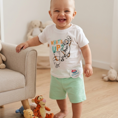 Bebé sorridente com camisola branca e calções verdes numa sala com peluches no chão.