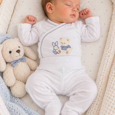 Bebé a dormir com fato branco e bordado de coelho e urso numa cama com edredão branco e peluche