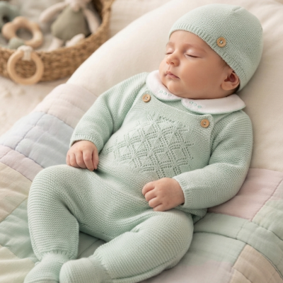 Bebé vestido com roupa verde-água em malha com detalhes e gorro, deitado numa manta acolchoada com brinquedos ao fundo.