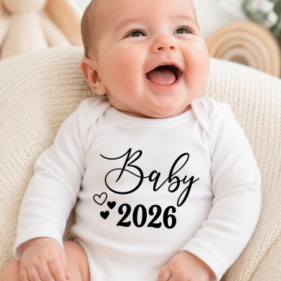Bebé sorridente com body branco de manga comprida com texto Baby 2026 preto, sentado numa almofada creme com brinquedo e planta ao fundo.