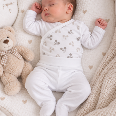 Bebé vestido com fato branco e padrão Mickey deitado em colchão com corações e ursinho de peluche ao lado