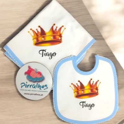 Conjunto de babete e fralda brancos com bordas azuis, coroa laranja e nome Tiago sobre mesa de madeira