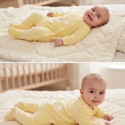 Bebé de macacão amarelo com ursinho deitado e de bruços numa cama clara com berço de madeira ao fundo