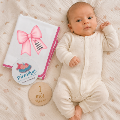 Bebé com fato creme numa manta decorada com estrelas e luas, ao lado de pano branco com laço rosa e texto, etiqueta da marca e placa de madeira com data