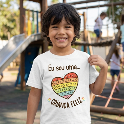 Camiseta branca infantil com estampa colorida de coração pop-it e texto em português usada por criança sorridente num parque
