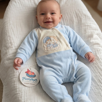 Bebé sorridente vestido com macacão azul e branco com bordado de ursinho, deitado numa manta branca com etiqueta pendurada.