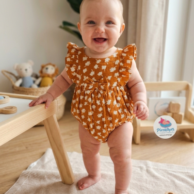 Body de bebé castanho alaranjado com flores brancas e laço, bebé sorridente em ambiente interior com tapete e brinquedos de madeira