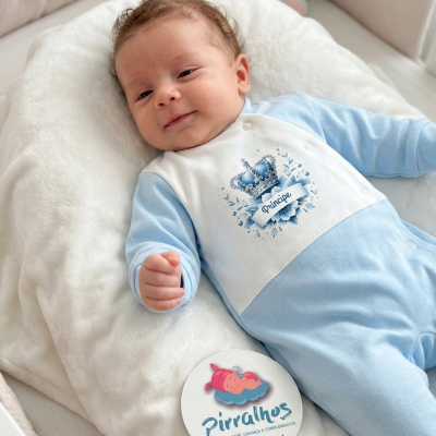 Bebé vestido com macacão azul claro e branco com estampa de coroa e texto Príncipe deitado em almofada branca felpuda com etiqueta redonda da marca Pirralhos