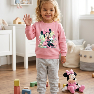 Criança com camisola rosa da Minnie Mouse e calças cinza em quarto infantil com brinquedos e peluche.