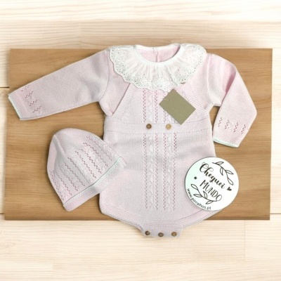 Body e gorro de bebé rosa clara em malha com colarinho de renda branca e botões de madeira, sobre tábua de madeira clara.