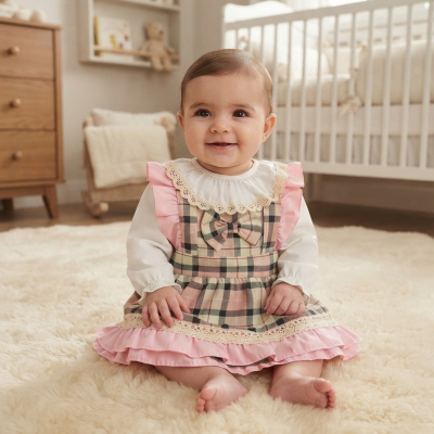 Bebé sentada com vestido xadrez rosa e bege com detalhes em rosa e renda creme, blusa branca de manga comprida, em tapete felpudo beige junto a berço branco.
