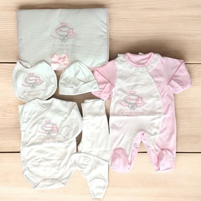 Conjunto de roupa de bebé rosa e branca com bordados, dispostos sobre superfície de madeira clara.