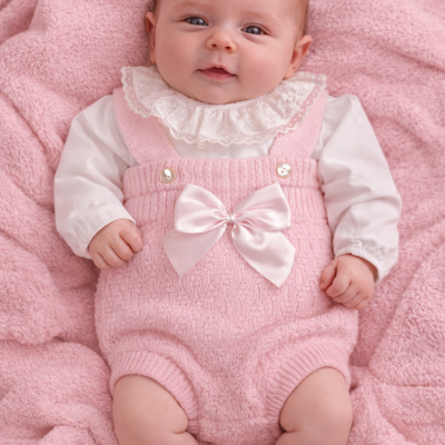 Bebé com roupa de malha rosa e camisa branca de gola rendada, sobre manta rosa felpuda.