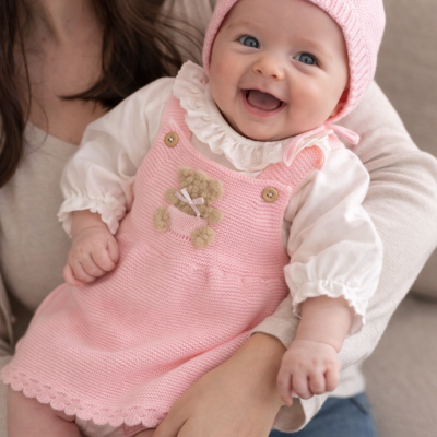 Bebé sorridente com vestido rosa de malha, camisola branca com folhos e gorro rosa, nos braços de uma mulher.
