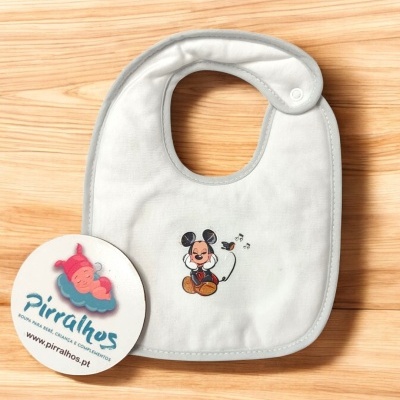 Babete para bebé branco com desenho do Mickey Mouse, sobre superfície de madeira clara com cartão promocional