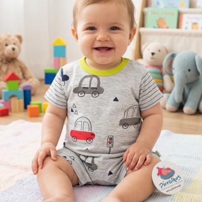 Bebé sorridente com macacão cinzento de carros sentado em manta colorida, quarto com brinquedos e peluches ao fundo