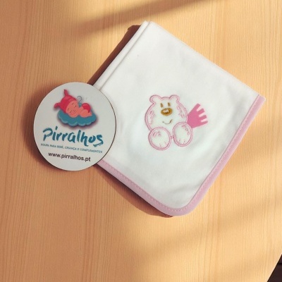 Fralda branca com borda rosa e desenho de ursinho, etiqueta Pirralhos no fundo de madeira