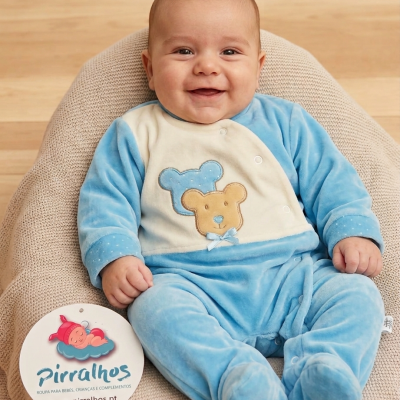 Bebé de macacão azul com ursinho bordado sentado em almofada bege, com etiqueta da marca Pirralhos