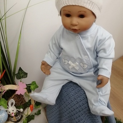 Boneco bebé com fato azul claro e gorro branco sentado sobre manta azul perto de plantas e brinquedos