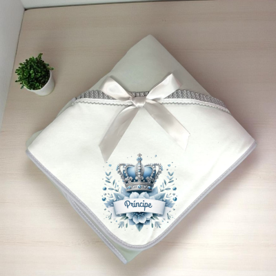 Manta branca com coroa azul e texto Príncipe em superfície de madeira com planta ao lado