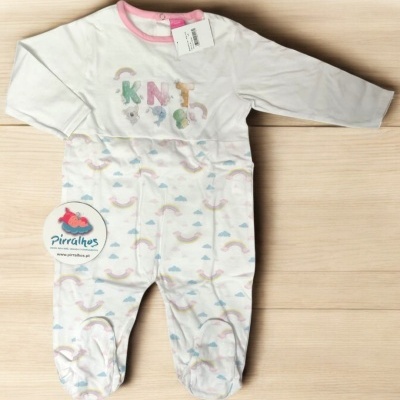 Pijama de bebé branco com detalhes em rosa e estampado de arco-íris, em superfície de madeira clara