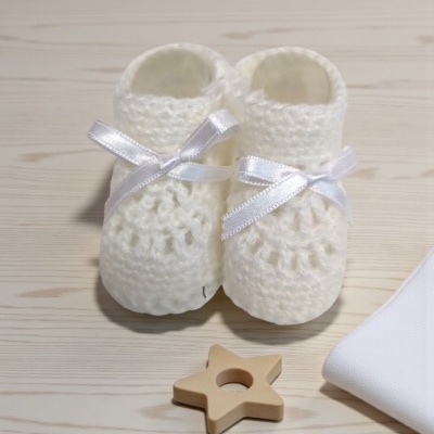 Pantufas brancas de bebé em malha grossa com laços brancos sobre mesa clara, estrela de madeira e tecido branco.