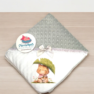 Manta de bebé branca e cinza com desenho de gatinho e laço, etiqueta Pirralhos, sobre superfície de madeira.