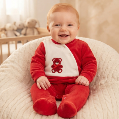 Bebé sorridente com macacão vermelho e branco com ursinho bordado sentado em almofada tricotada bege