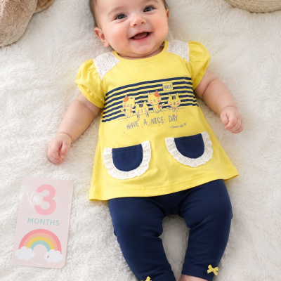 Bebé com túnica amarela e leggings azuis deitado em tecido branco, a sorrir, com cartão rosa com 3 MONTHS e arco-íris ao lado.