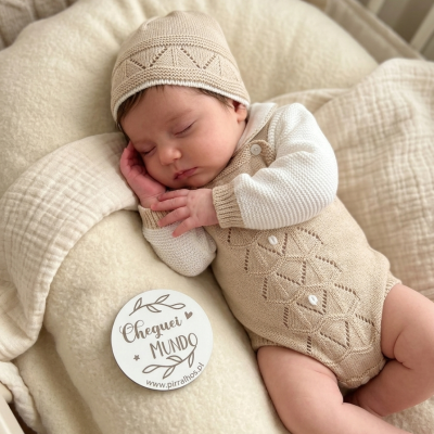 Bebé a dormir com roupa de malha bege e branca num berço com almofada creme e placa decorativa.