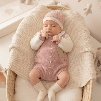 Bebé com macacão rosa e gorro de malha a dormir numa cesta com manta creme.