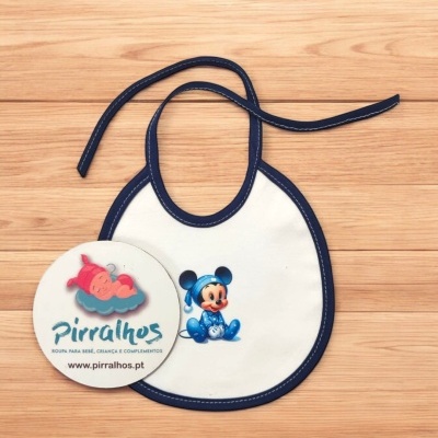 Babete de bebé branco com borda azul escura e padrão de rato azul, com cartão redondo promocional ao lado, sobre superfície de madeira clara