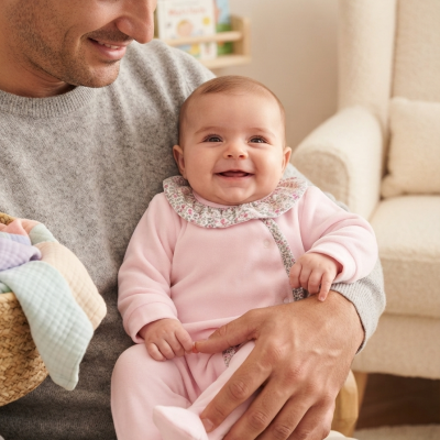 Bebé vestido de rosa com gola floral, sorridente no colo de um adulto num ambiente de casa