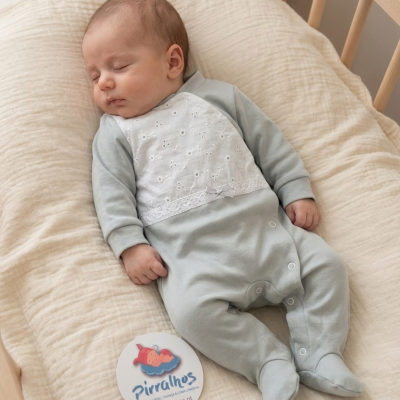 Bebé adormecido com fato cinzento com bordado branco deitado em almofada creme dentro de berço de madeira