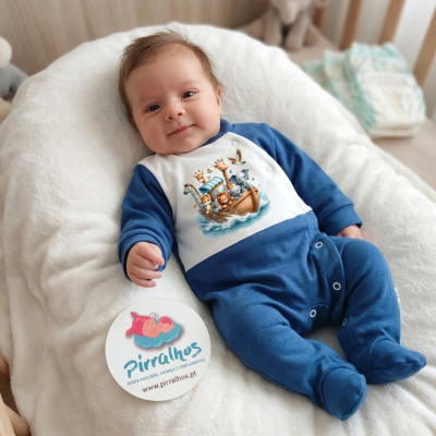 Bebé em fato de pijama azul e branco com estampado de animais em barco, deitado numa almofada branca num berço com peluche e etiqueta promocional.