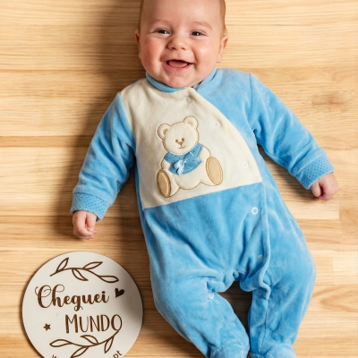 Bebé com fato azul claro com urso bordado a sorrir deitado no chão de madeira ao lado de placa branca com texto.