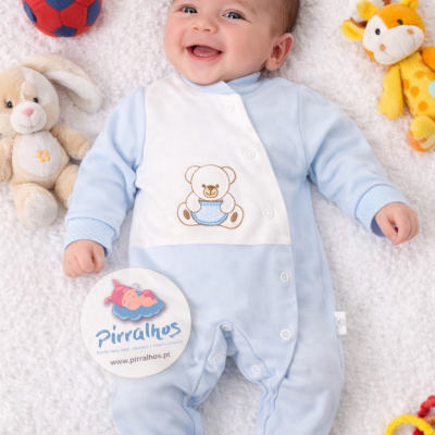 Bebé sorridente deitado numa manta branca com roupa azul clara com ursinho bordado e brinquedos ao redor