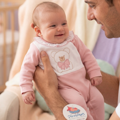 Bebé sorridente com macacão rosa com ursinho bordado segurado por adulto num ambiente interior.