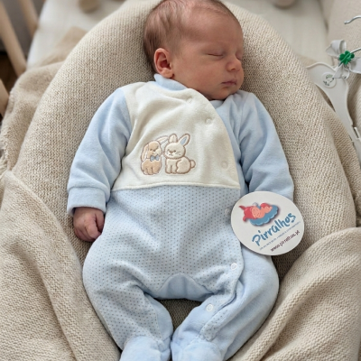 Macacão de bebé azul e branco com coelhinhos bordados, tecido macio, em almofada bege felpuda