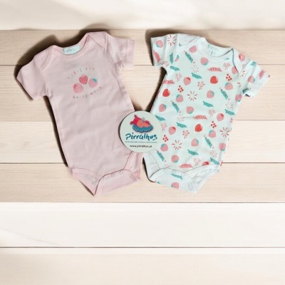 Dois bodies para bebé, um cor-de-rosa e um branco com padrão de morangos, sobre madeira clara com etiqueta 'Pirralhos'.
