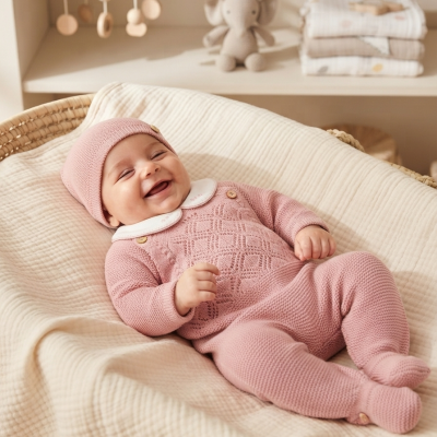 Bebé vestido com fato de malha cor-de-rosa com gola branca, deitado em cesta com manta creme.