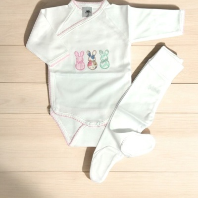 Conjunto de body branco com três coelhinhos bordados e meias brancas, em chão de madeira clara.