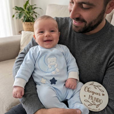 Bebé com fato azul claro com estampado de ursos, lua e estrelas sentado nos braços de adulto com camisola cinzenta em sala com sofá e planta.