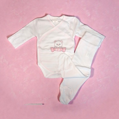 Conjunto branco de roupa de bebé com bordado de urso e doce em fundo rosa.