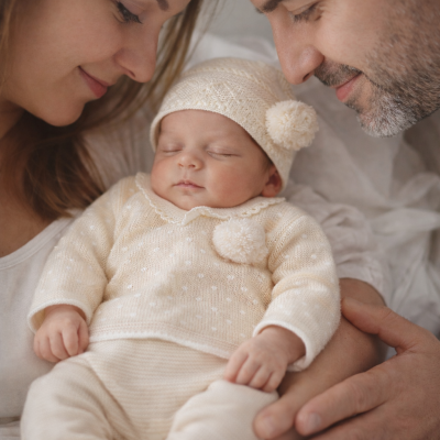 Bebé a dormir com roupa de malha creme com pompons, apoiado pelos pais.