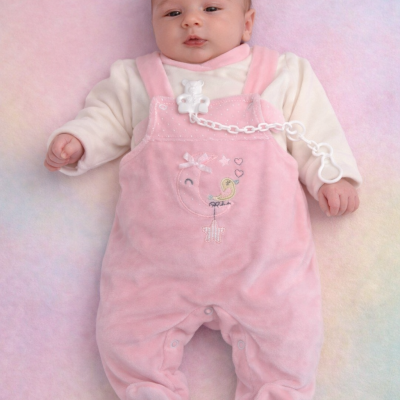 Bebé vestido com macacão rosa aveludado com bordado, chupeta presa com clip branco e cadeia, deitado em superfície macia colorida.