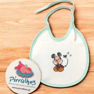 Babete branco com borda verde e estampa de Mickey Mouse ao lado de cartão redondo com logo e texto Pirralhos.
